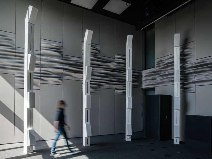 A tall grey room lined with undulating printed wall panels, with white loudspeaker columns and a person walking past