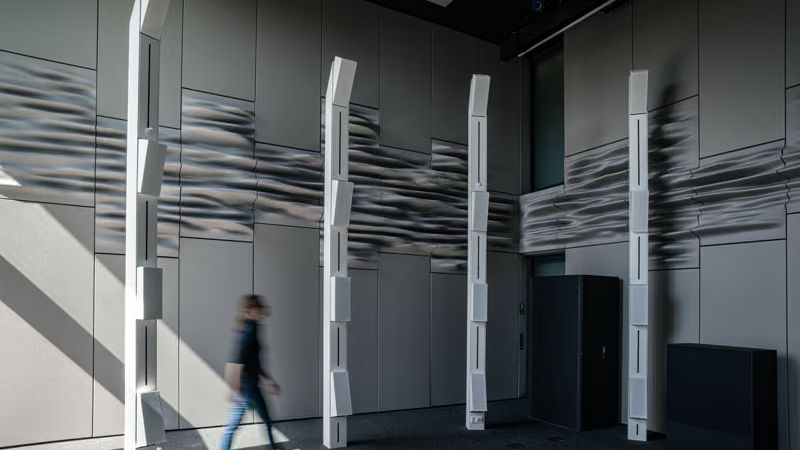 A tall grey room lined with undulating printed wall panels, with white loudspeaker columns and a person walking past
