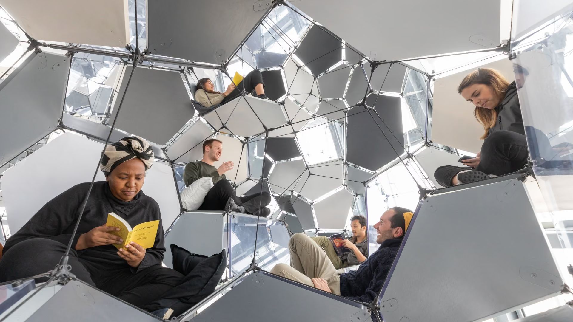 Visitors sitting and reading among suspended reflective polygonal panels connected by tensioned cables