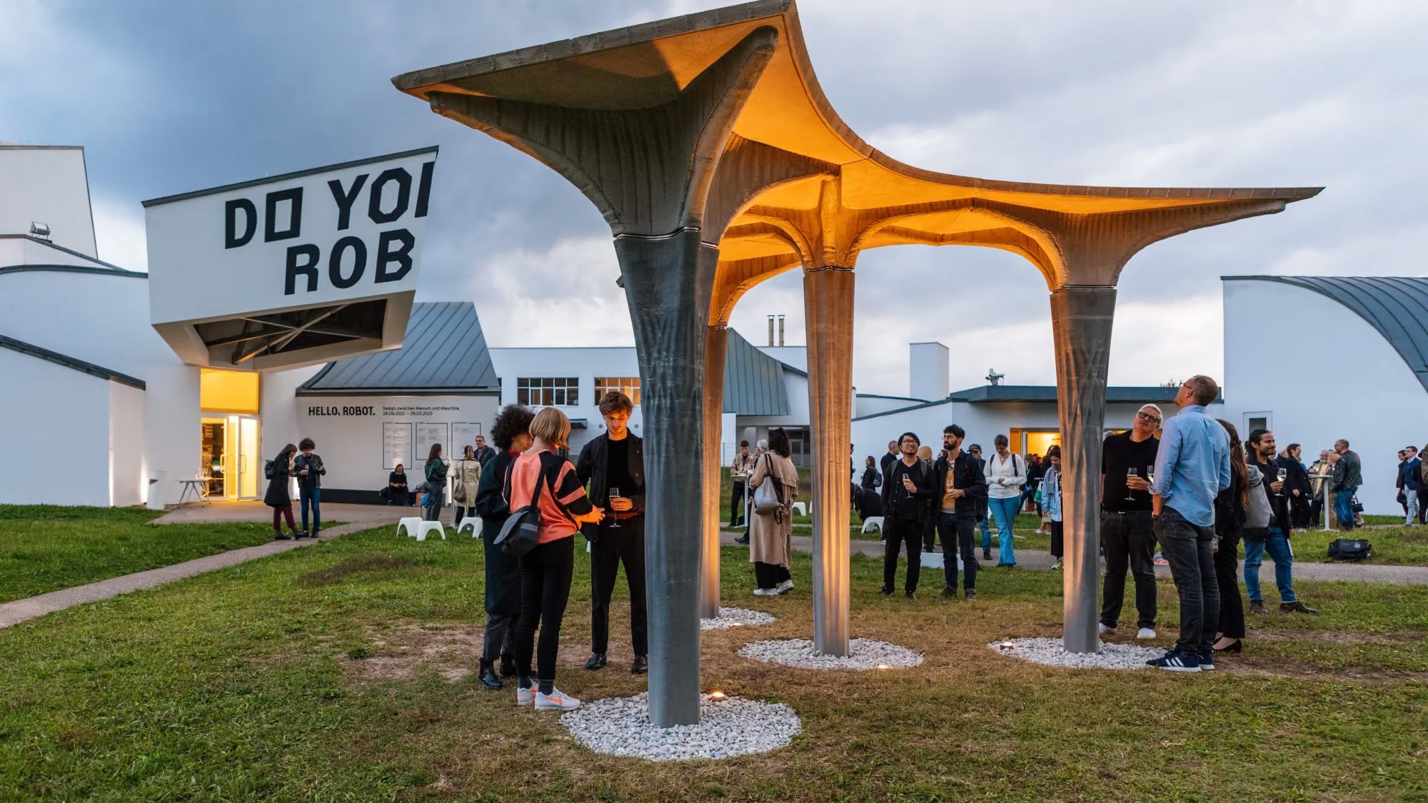Visitors at dusk around a pavilion of slender concrete columns and thin slabs lit warm from below, in front of the Vitra Design Museum