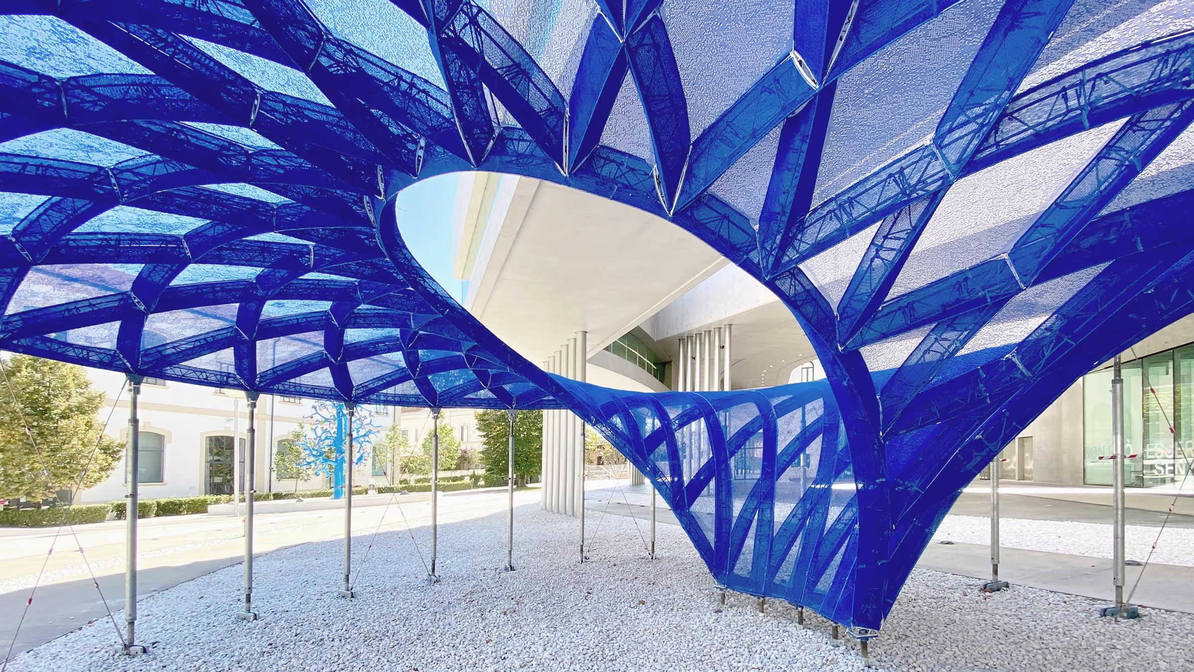 A blue knitted ribbed shell with a large oculus, raised on slender steel props outside a museum