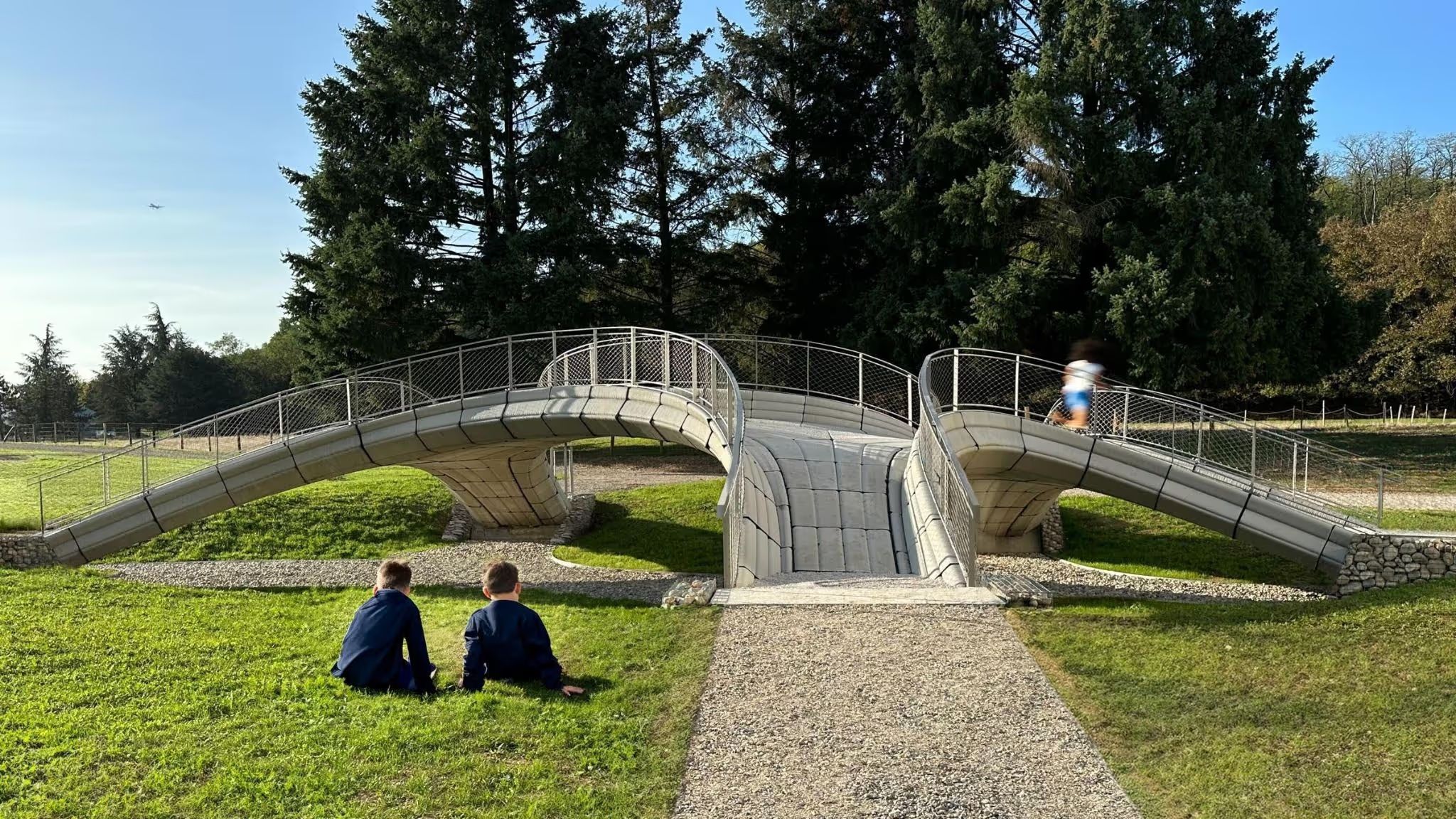 Two people sitting on the grass in front of a curving concrete footbridge with steel railings