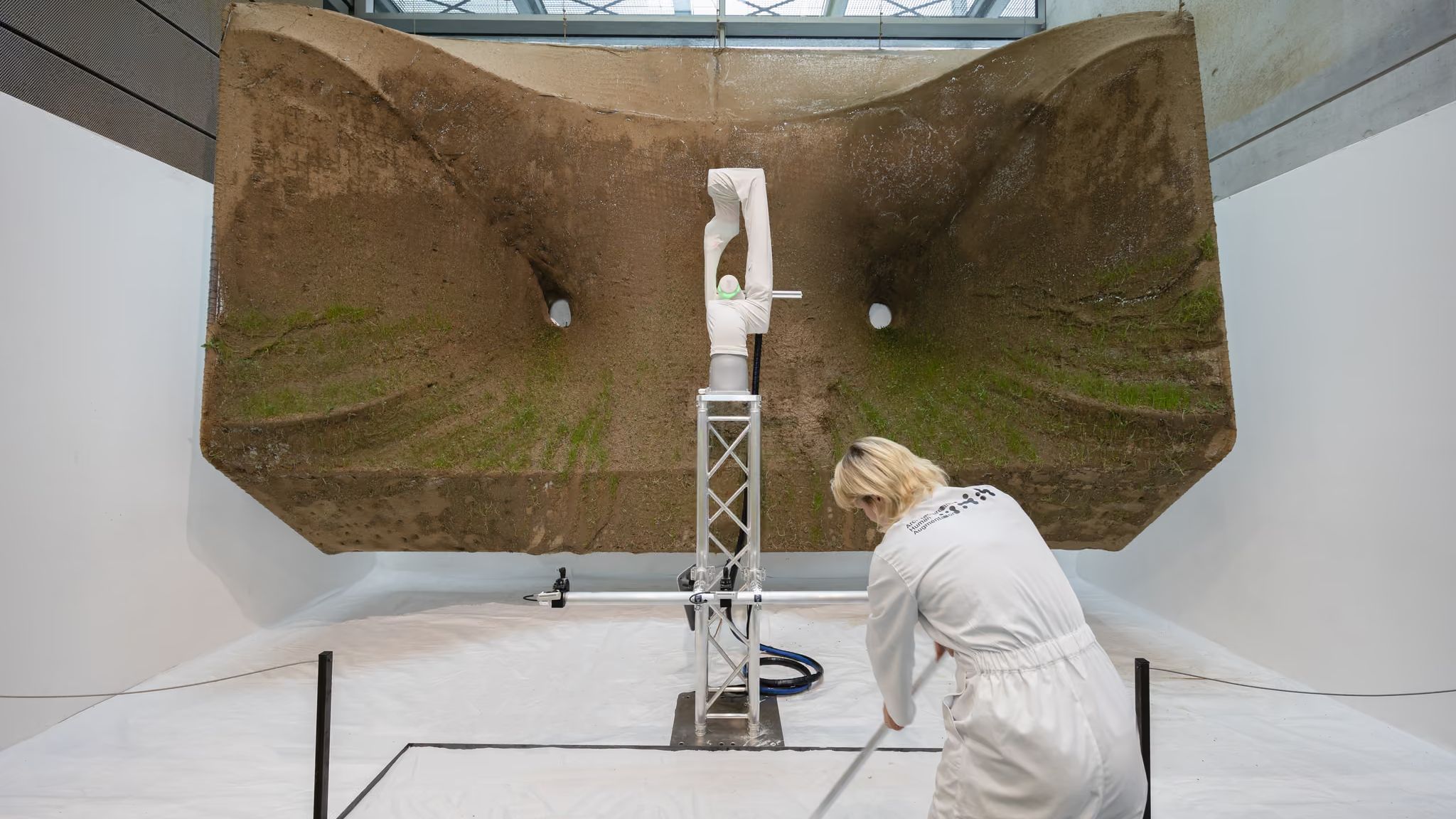 A robot arm on a truss in front of a large suspended net sprayed with soil and sprouting grass, while a person in a white overall draws in sand