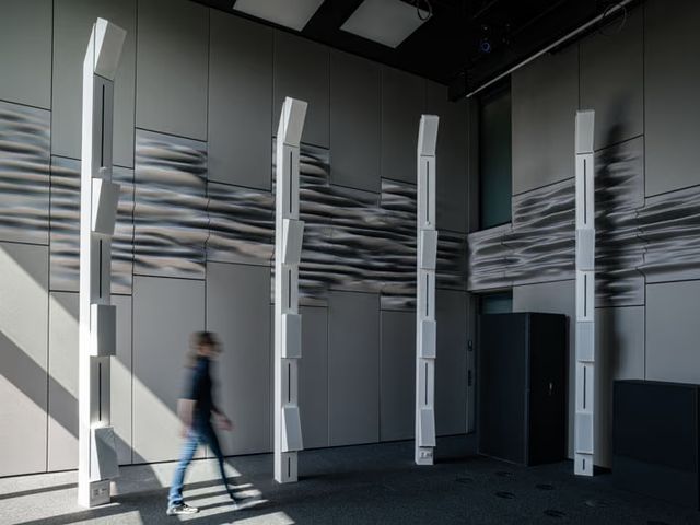 A tall grey room lined with undulating printed wall panels, with white loudspeaker columns and a person walking past