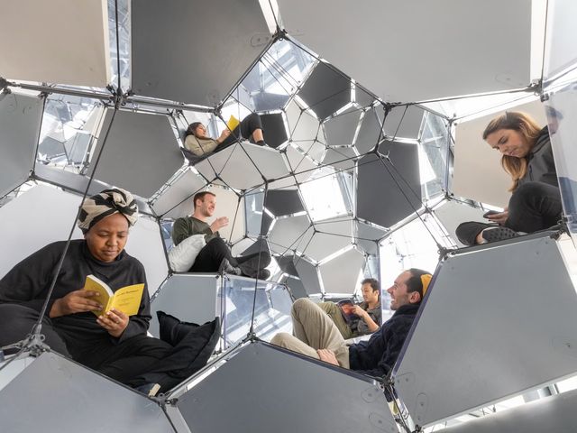 Visitors sitting and reading among suspended reflective polygonal panels connected by tensioned cables