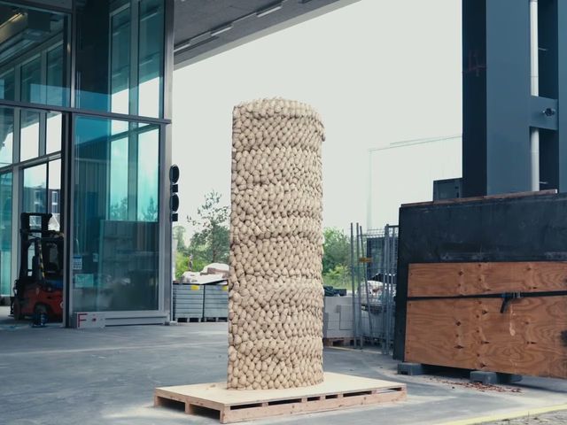 A column built from thousands of compressed earth portions standing on a pallet outside a laboratory hall