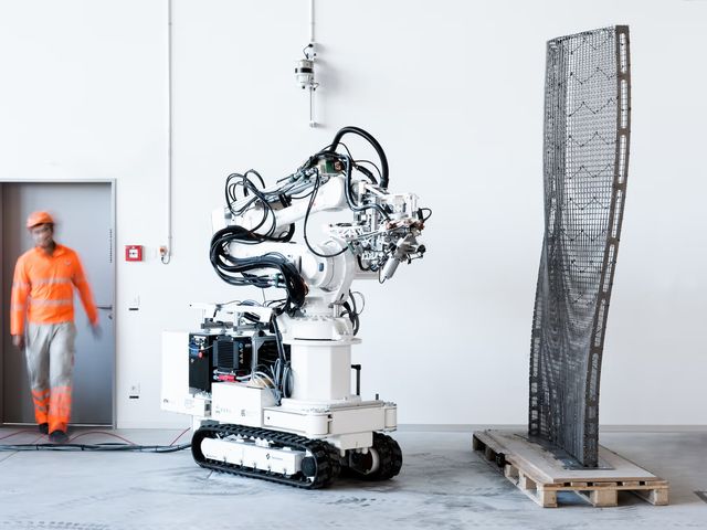 A worker walking past a tracked robot that stands beside a tall, twisting steel mesh it has fabricated