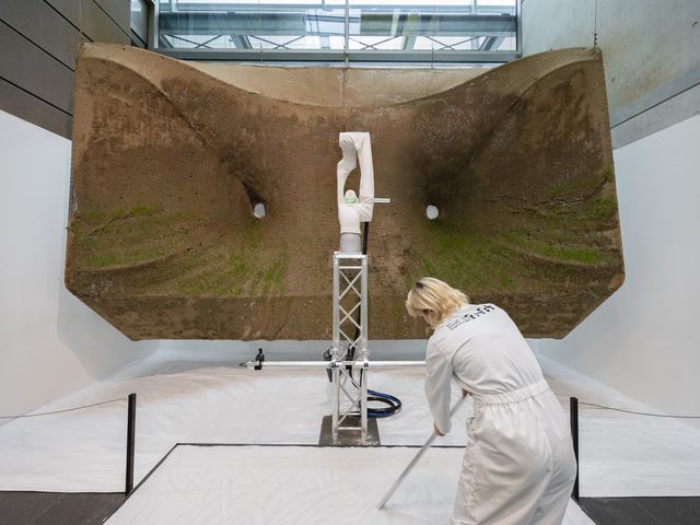 A robot arm on a truss in front of a large suspended net sprayed with soil and sprouting grass, while a person in a white overall draws in sand
