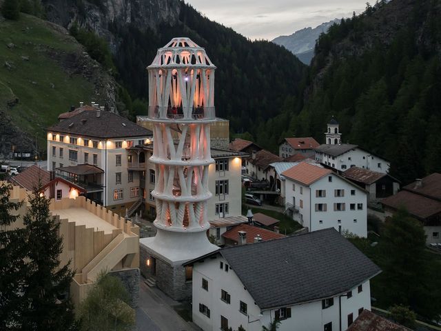 A tall white tower of branching printed concrete columns, lit from within at dusk, rising between houses in a mountain village